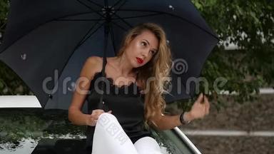 美丽的年轻女人，留着长长的金发，坐在白色汽车的引擎盖上，带着雨伞，在雨天。 慢慢慢慢